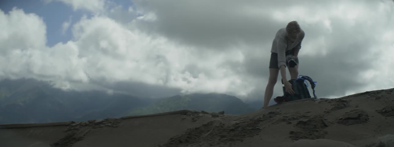 Photographer wearing shorts takes out his equipment while standing on a dirt hill on a cloudy day