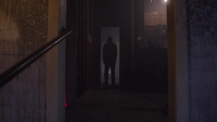 A static shot captures the dark silhouette of a person standing alone in a dimly lit narrow hallway or doorway the figure is facing the camera giving the scene an ominous mysterious or isolating atmosphere