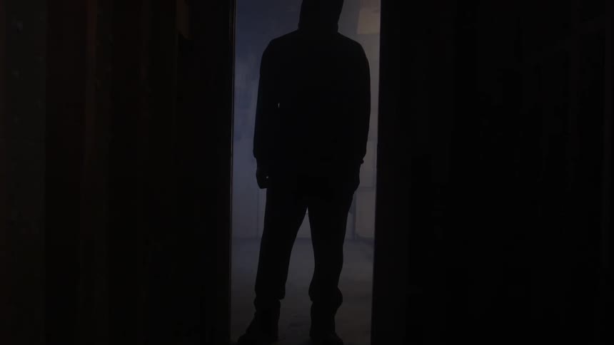 Man wearing a hoodie stands silhouetted in a door leading to a dark hallway