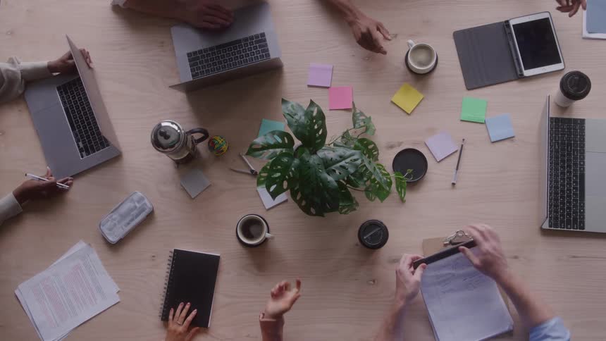 This is a highangle overhead shot of several individuals gathered around a light wooden table during a meeting the table is cluttered with open laptops papers coffee cups and a central potted plant with hands moving items around and interacting wit