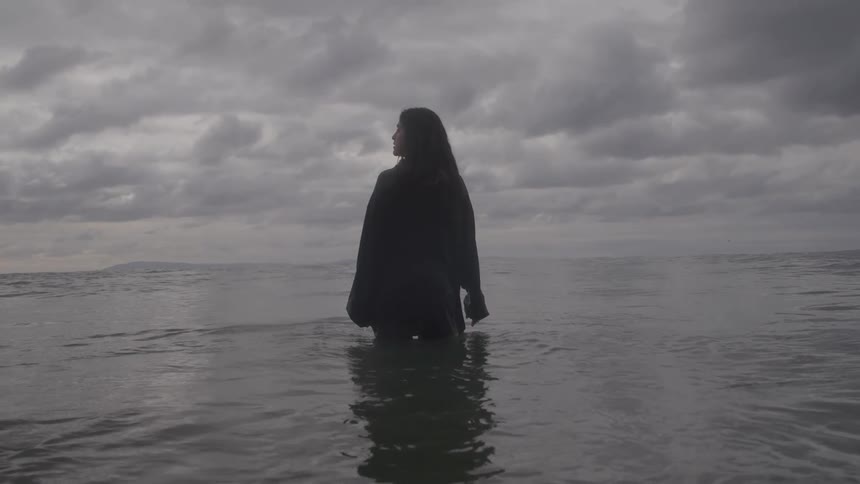 A person with long dark hair is standing waistdeep in the ocean or a large body of water seen from behind and slightly to the side the scene has a somber monochromatic feel with dark water and a heavy overcast sky filling the background