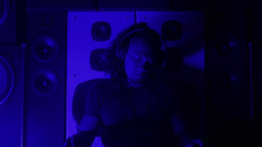 A tattooed man wearing a dark shirt and headphones sits centered before a wall of large audio speakers bathed in flashing blue and red club lighting
