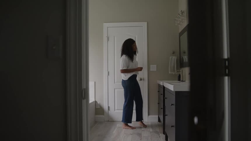 The video shows a woman standing in a bathroom or dressing area with a white buttondown shirt tucked into dark blue pantsshe is adjusting her shirt or getting dressed with her face partially obscured by shadow as she looks down at her hands