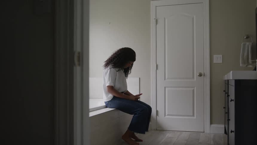 A shot from a doorway shows a young woman with curly hair sitting on the edge of a bathtub in a bright modern bathroom she is initially looking up then looks down and focuses on something she is holding in her hands possibly a phone
