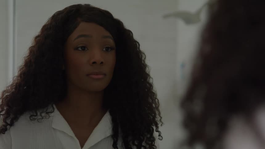 The video is a closeup shot of a young black womans face with long dark curly hair looking downwardshe is reflected in a mirror directly in front of her which suggests a moment of introspection or personal preparation
