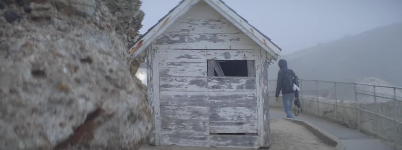 A person in a hoodie and backpack walks past a small wooden shed