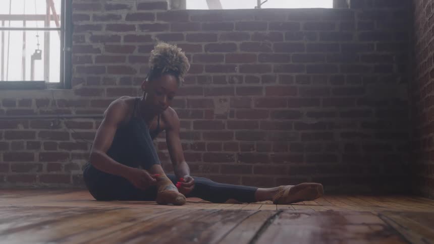 A female dancer is sitting on a wooden floor in a studio facing a red brick wall with a window she is wearing dark dancewear and is adjusting the straps of her ballet or jazz shoes
