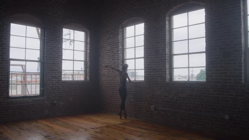 A ballet dancer in black fitted clothing performs alone at midday on a wooden floor inside a spacious empty room