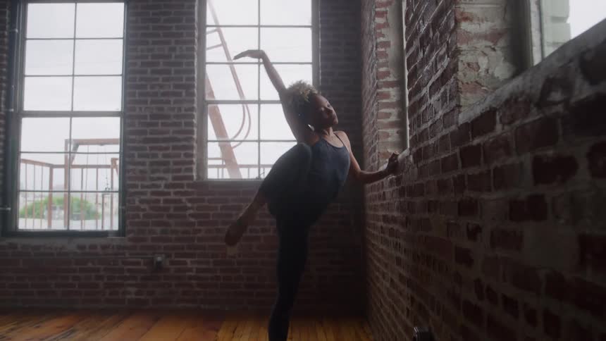 A black female ballet dancer in black tight clothing and ballet shoes stretches beside a wall at midday inside a large room with a wooden floor