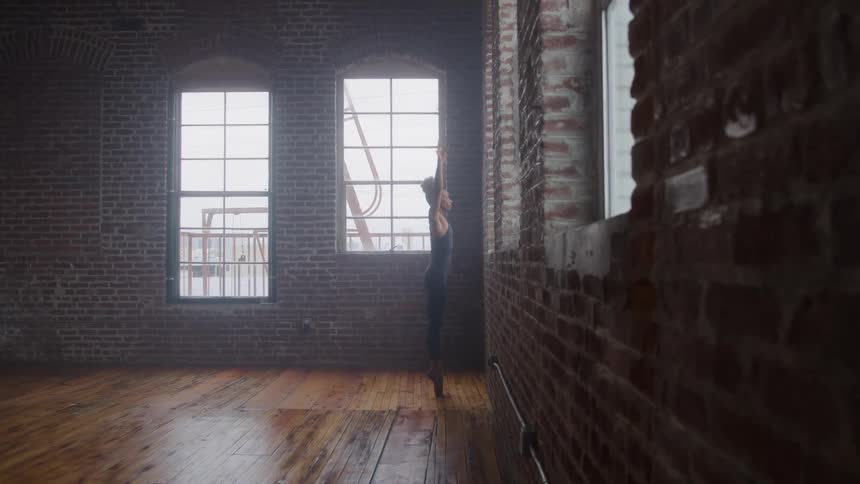 A female ballet dancer stretches her arms and body beside a brick wall at midday in a spacious room with a wooden floor