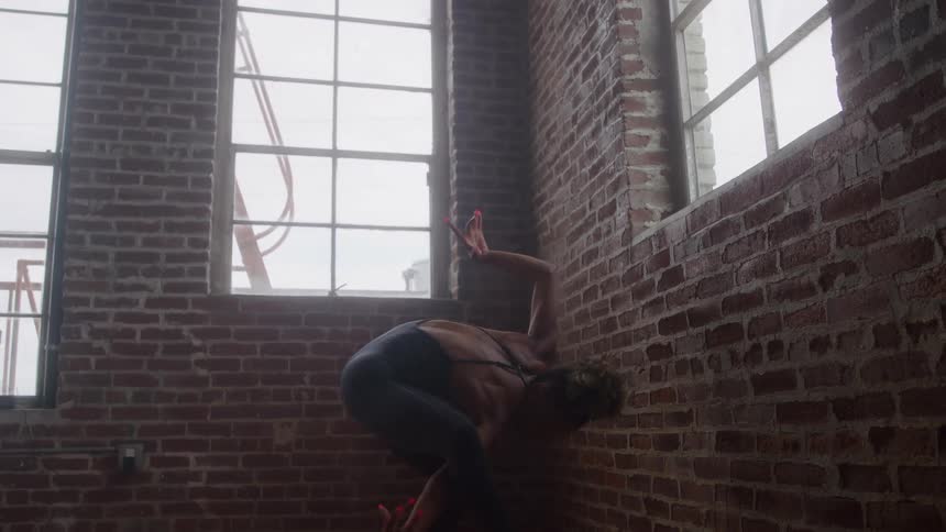Woman wearing a leotard dances in the corner of a brick walled room