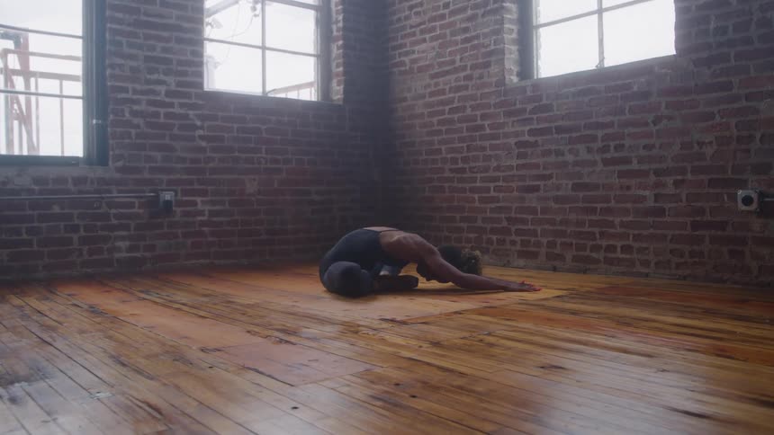 Woman performs balasana pose on mat in spacious room with wooden floor and sunlit windows