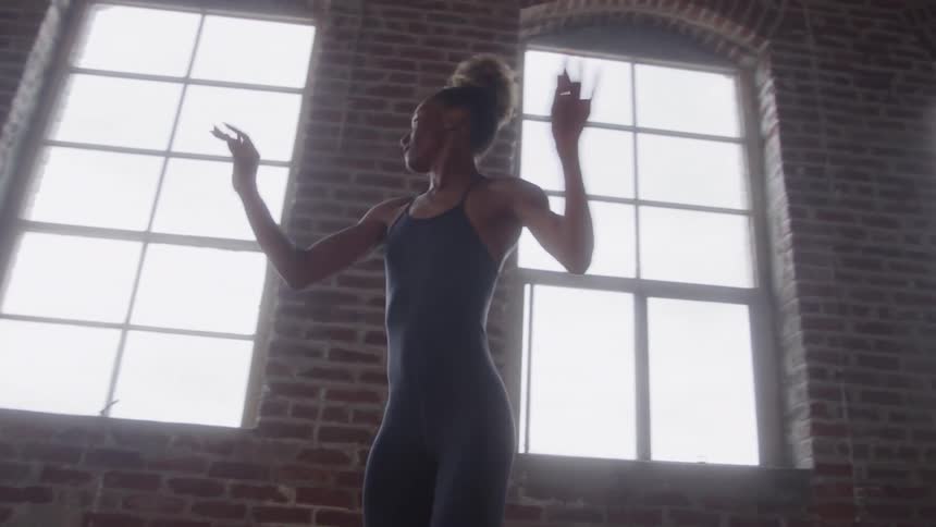 A young woman ballet dancer practices her moves alone at midday in an empty room with multiple windows