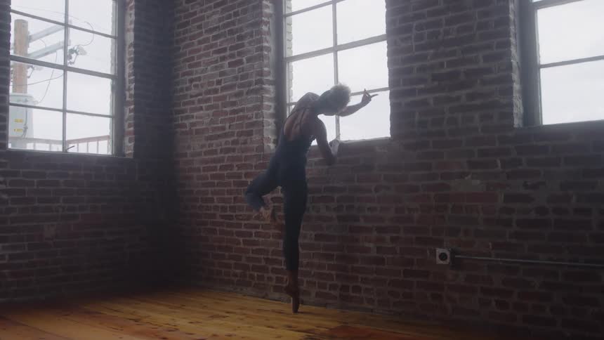 A young woman in black clothing and ballet shoes stands stretching near a window at midday gazing outside from a room with a wooden floor
