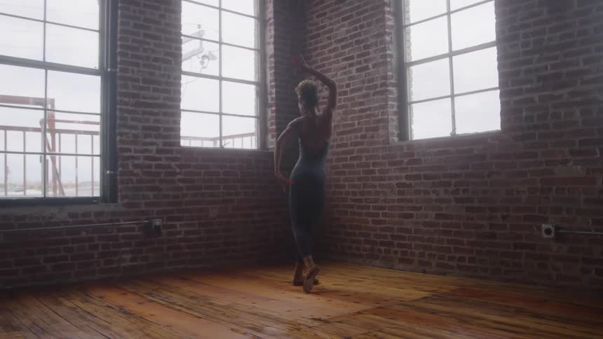 A young woman dances and practices ballet inside a spacious room with a wooden floor and multiple windows at midday