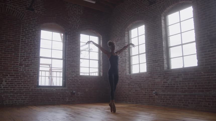 A young female ballet dancer balances on her toes with her hands slowly raised during a midday training session in a spacious room with a wooden floor