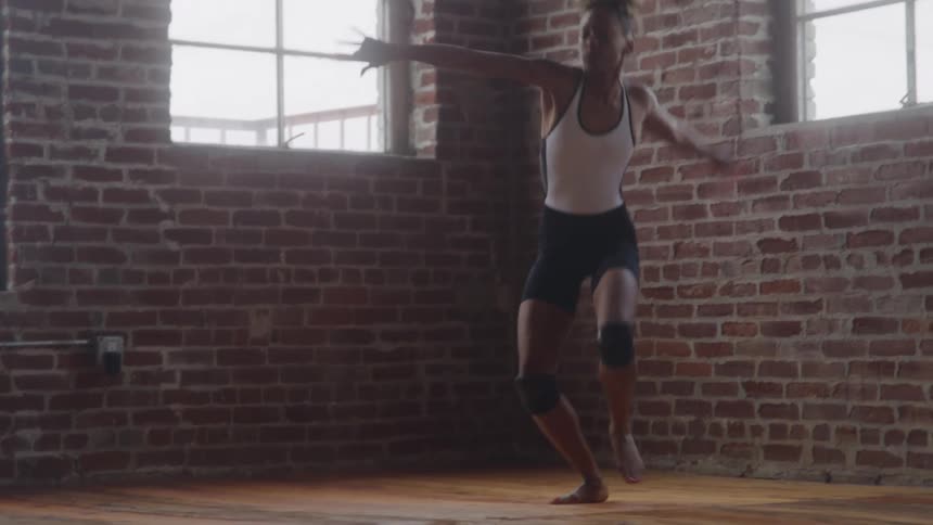 A young woman in activewear performs acrobatic dance moves on a wooden floor at midday