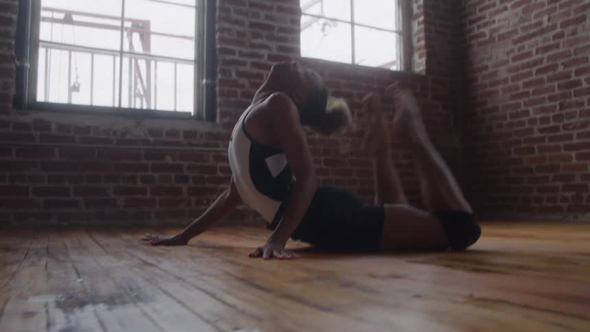 A young woman in activewear performs a yoga dance exercise on a wooden floor at midday