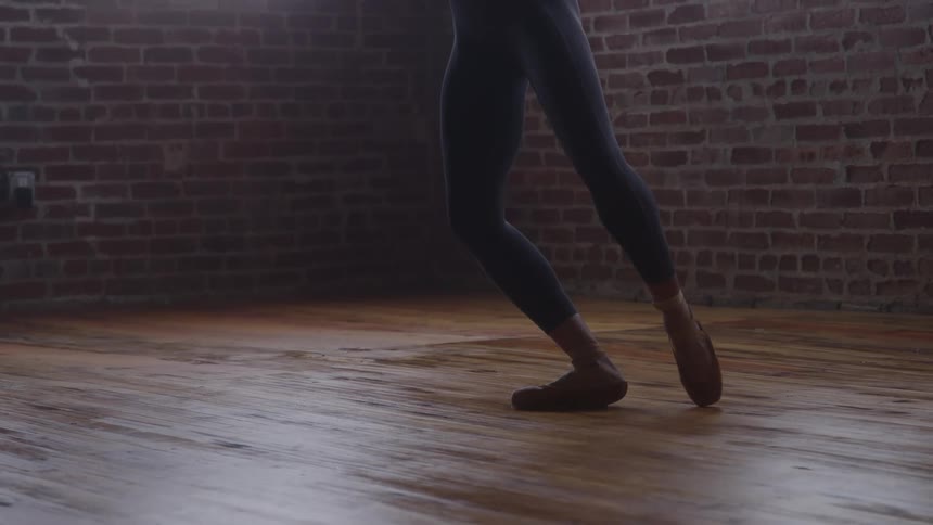 Person in ballet shoes performs pointe position in studio with wooden floor and exposed brick wall