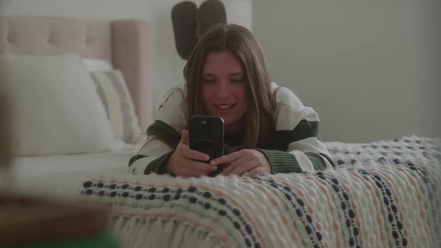 A young woman lies on her bed in her bedroom at midday smiling as she uses her phone