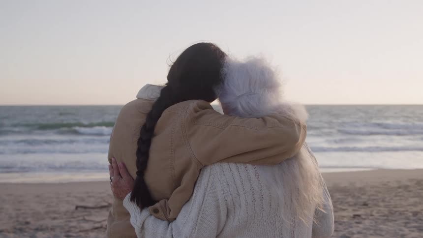 An elderly woman with long grey hair hugs a young woman as they stand together on the beach sand at midday both wearing sweatshirts and watching the ocean waves