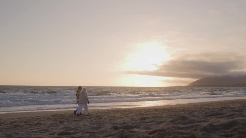 Two women one an older woman with long grey hair walk together along the shoreline of a sandy beach as gentle waves roll in during a bright hazy sunset