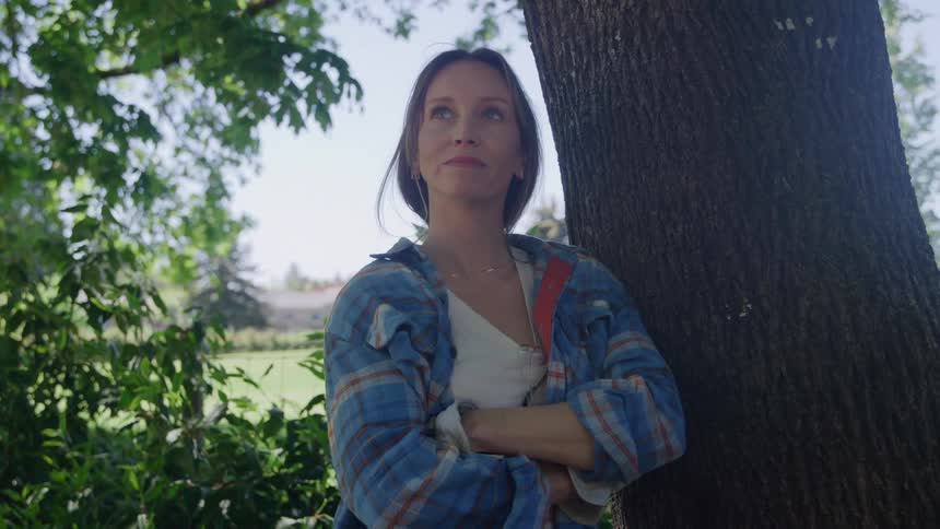 A woman with long brown hair stands outdoors leaning her back against a tree trunk with arms crossed over a white top and plaid shirt looking upward past green leaves under bright sun