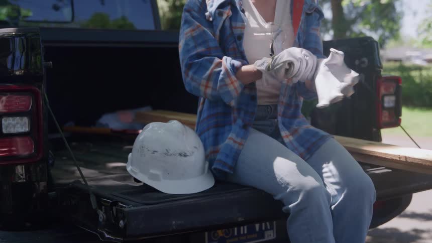 A woman sits on a truck tailgate putting on work gloves in daylight