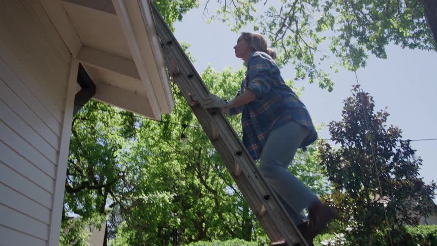 A woman stands on a ladder repairing the roof of a house among green trees
