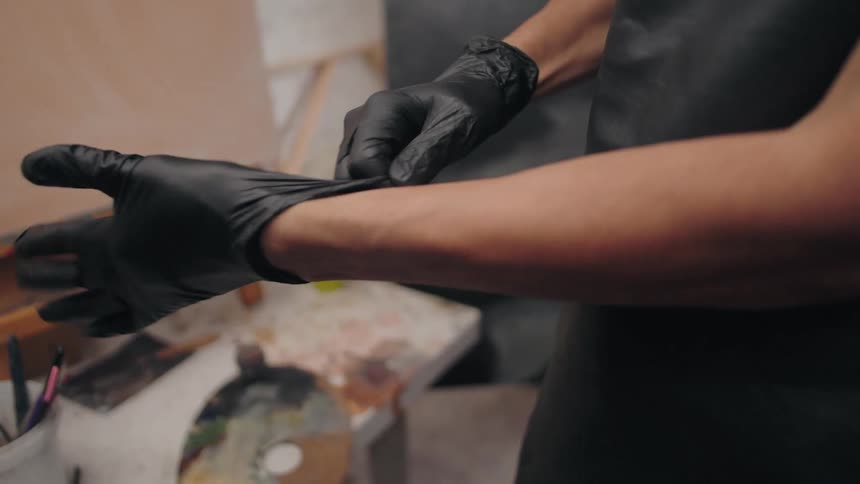A person wearing a black apron stands in a studio pulling a black glove onto their right hand with the left gloved hand preparing to begin work at a table with art supplies visible