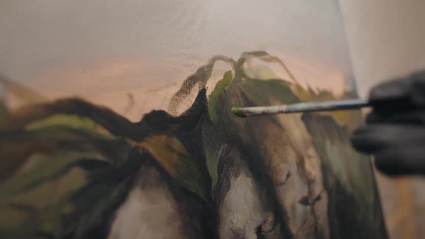 An artist wearing a black glove carefully applies a dab of green paint with a fine brush to a detailed oil painting of a mountain landscape on canvas