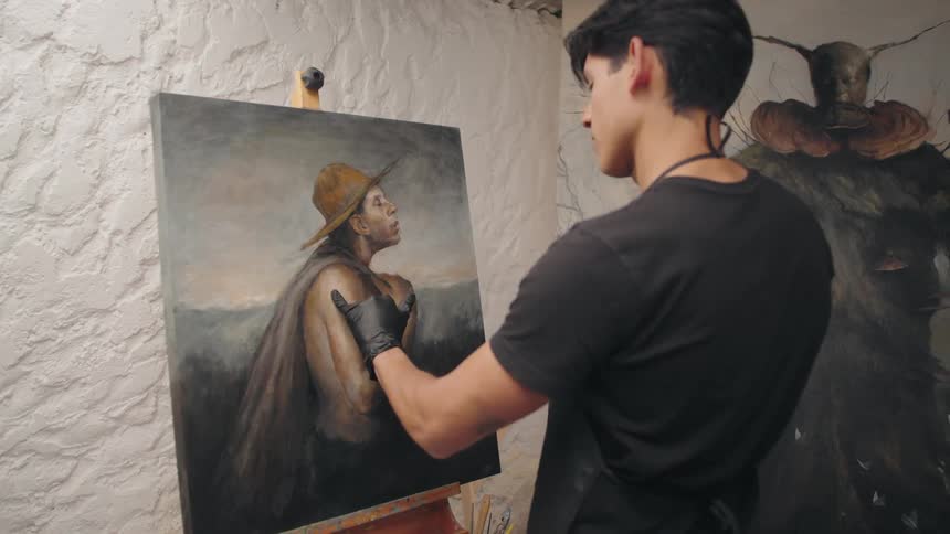 A young male artist in a black shirt and apron stands in his studio intently working on a large moody oil painting of a figure in a hat