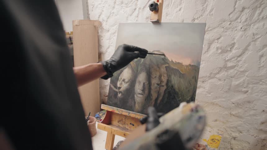 An artist in black gloves carefully adds details to a surreal painting of faces in a landscape