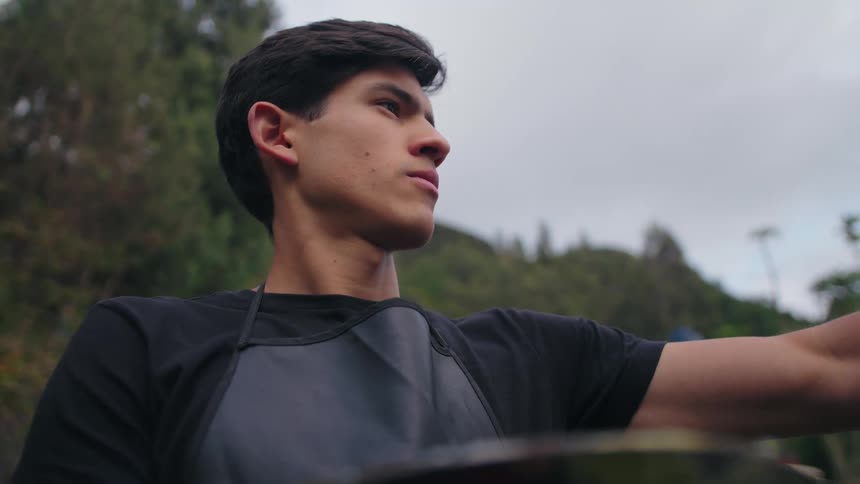 A young man wearing a black shirt and a dark apron is outdoors with a blurred green background possibly preparing to cook or paint he is holding a large bowl or pan and looking away with a determined expressioncan i describe something el