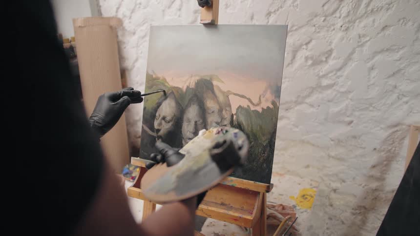 A closeup shot focuses on an artists hands wearing black gloves meticulously painting on a canvas set on an easel the artist holds a palette with various colors in one hand while applying paint to a piece with abstract figures