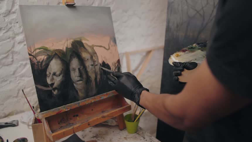 An artist in black gloves paints a portrait on canvas with a brush focused and steady inside the studio