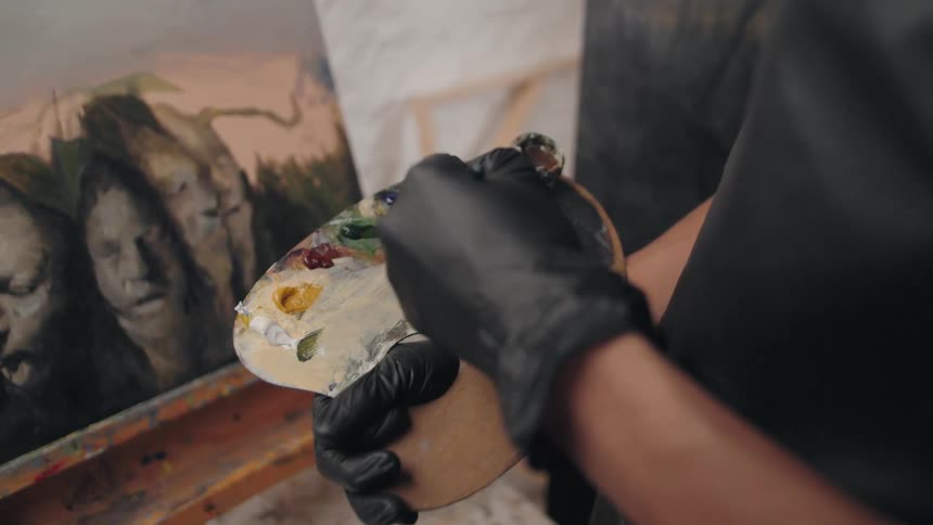 An artist wearing black gloves expertly mixes oil paints on a wooden palette with a fine brush in front of a nearly finished portrait on an easel