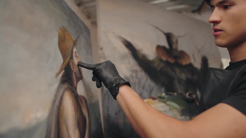 A painter moves a gloved finger to apply paint directly onto a canvas a palette rests in the hand and another canvas with a darker subject stands in the background
