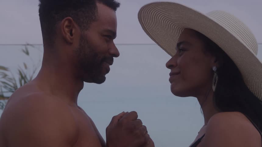 A closeup side profile shot of a man and a woman holding hands and gazing affectionately at one another the man is shirtless and the woman is wearing a dark top and a large straw sun hat
