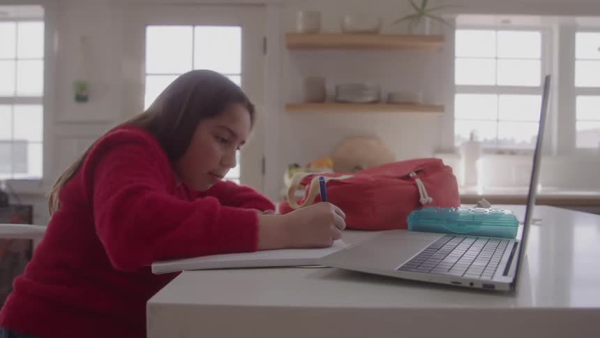 A young girl in a red sweater is sitting at a kitchen counter focusing intently on writing in a notebook a partially open laptop and some school supplies sit beside her