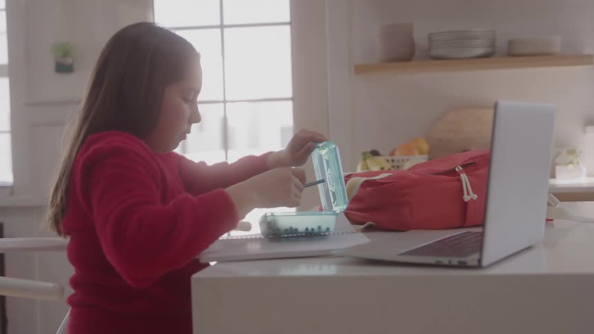 A young woman with long hair wearing a red sweater sits at a kitchen counter with a laptop and a red backpack taking a blue pencil case out to begin her homework