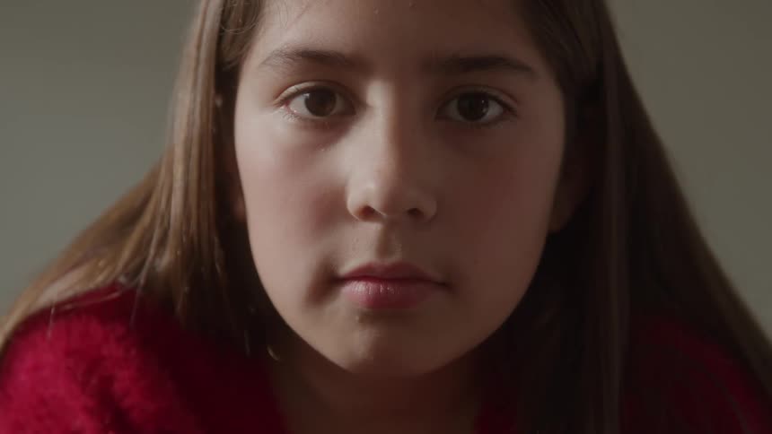 A tight closeup focuses on the face of a young person with long brown hair looking straight into the camera they maintain a neutral serious expression throughout the short clip