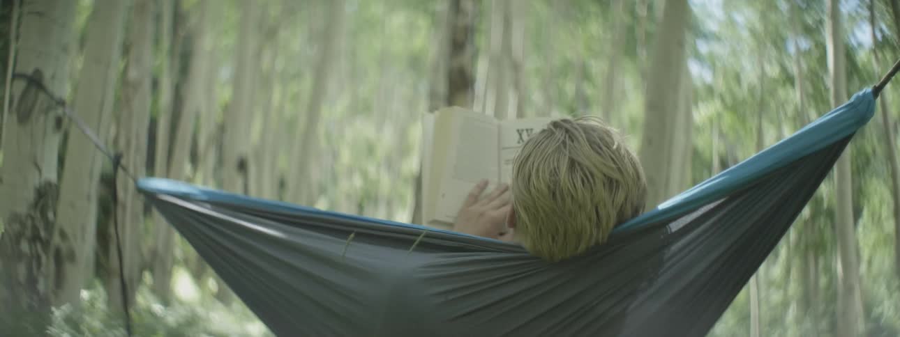 Man laying in a hammock in the woods reading a book
