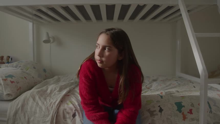 A teenage girl sits alone on the bottom bunk in her bedroom at midday wearing a red sweatshirt and appearing quietly thoughtful in the calm light of the room