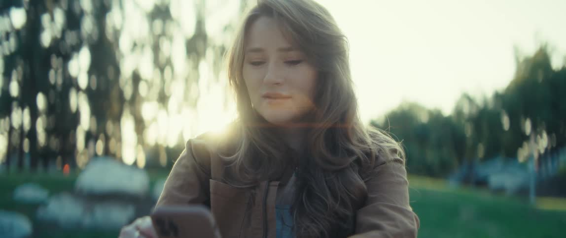 A woman smiles softly at her phone while sitting in a park at sunset
