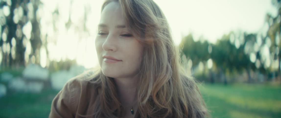 The video is a closeup shot of a young womans face outdoors lit by warm sunlight she has long light brown hair and is wearing a brown jacket over a gray shirt appearing to be writing or drawing something out of frame