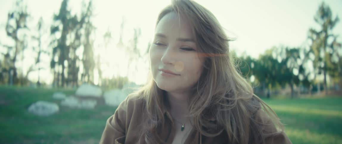 A woman with long sunlit hair sits outdoors at a table using her smartphone in landscape mode she appears to be watching or recording something adjusting her hair as she looks at the screen
