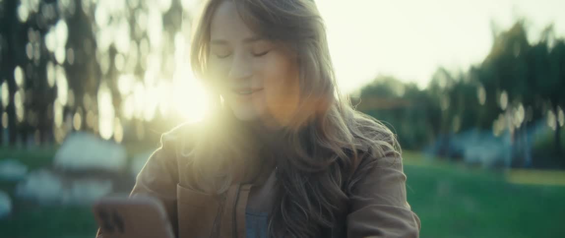 A woman looks down at a mobile phone she holds with both hands smiles and laughs with sunlight creating a bright lens flare around her in an outdoor setting