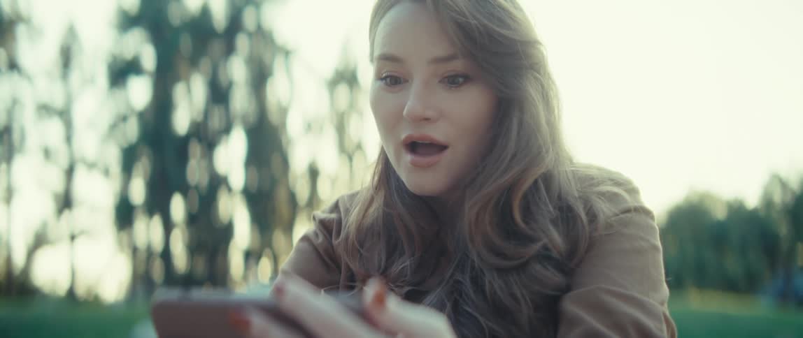 A young woman in a brown jacket is sitting outdoors examining a small white card she has a pensive expression as she looks at the item in her hands
