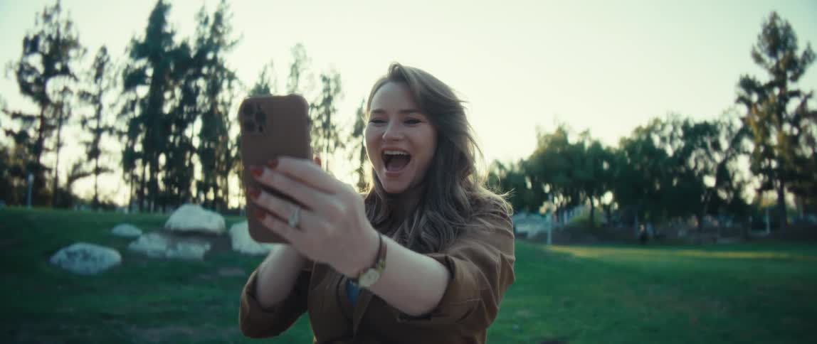 A woman sitting outdoors at a table with sunglasses resting near her is engaged with her smartphoneshe starts by looking down at the screen then lifts the phone as if on a video call or recording a video message smiling and speaking excitedly